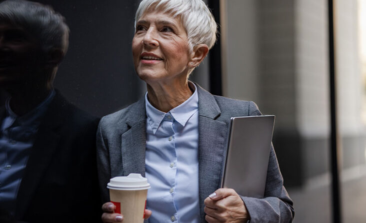 A woman holds a folder and cup of coffee as she heads to work, successfully balancing her career with care for aging parents.